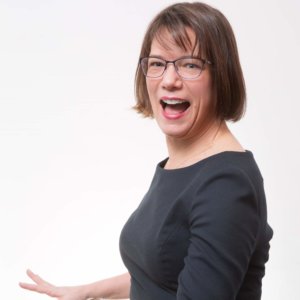 Professional headshot of smiling blonde woman in black blazer sitting in modern gray chair with laptop
